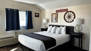 A neatly arranged hotel room with a large bed, two bedside tables with lamps, a heater, and a sign instructing guests to remove their shoes before bed. - Cabin 26 - Coyboy Corner (Helena)