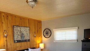 Un dormitorio con pared de paneles de madera, una cama con ropa de cama blanca, una mesita de noche con lámpara, un reloj de pared y una ventana con persianas.