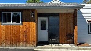 Un bâtiment d’un seul étage, avec une façade en bois, une porte blanche et deux chaises en métal à l’extérieur.