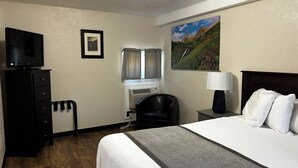 Une chambre d’hôtel avec un lit, un bureau avec une lampe, une télévision, une chaise, une fenêtre avec des rideaux et un tableau au mur.