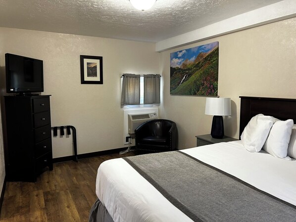 Une chambre d’hôtel avec un lit, un bureau avec une lampe, une télévision, une chaise, une fenêtre avec des rideaux et un tableau au mur.