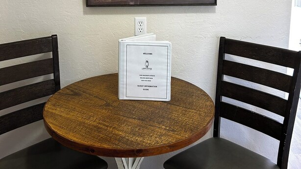 Une table ronde en bois, deux chaises noires, un tableau encadré représentant des fleurs accroché au mur et un panneau d’accueil avec des informations pour les clients.