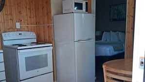 Une cuisine compacte équipée d’une cuisinière, d’un four, d’un réfrigérateur et d’un four à micro-ondes. La pièce adjacente contient une table en bois et un lit.