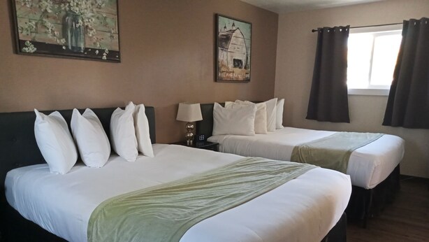 Une chambre d’hôtel avec deux lits, du linge de lit blanc, un plaid vert, une table de chevet avec une lampe et deux tableaux au mur.
