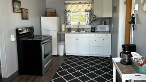 Cocina compacta con estufa, horno, refrigerador, microondas y lavaplatos. Hay una ventana con cortinas estampadas, un cubo de basura pequeño y una cafetera sobre la encimera.