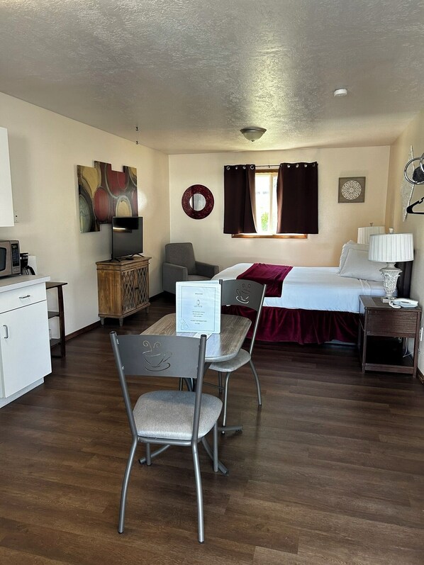 A compact hotel room with a bed, a desk with a chair, a small kitchenette, and a dining area. - Suite 33 - Queen Suite W/ Walk-In Shower (Helena)