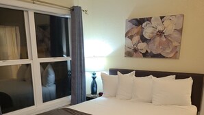 A hotel room with a large bed, a window with curtains, a painting on the wall, and a nightstand with a lamp. - Suite 34- Victorian Suite (Helena)
