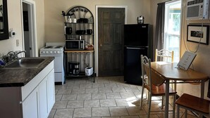 A compact kitchen with a stove, microwave, and refrigerator, a dining table with chairs, and a window with an air conditioner. - Suite 34- Victorian Suite (Helena)