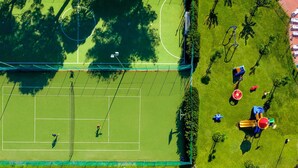 Vista aérea de una cancha de tenis con red, rodeada de árboles y un parque infantil con columpios y toboganes.