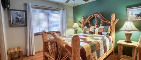 Una cama de madera con colcha de cuadros, un ventilador de techo, una ventana con persianas y un pequeño taburete de madera.