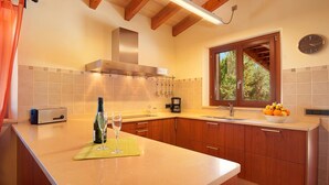 A kitchen with wooden cabinets, a built-in oven, a coffee maker, and a window with a view of trees. - La Salve d'alt villa with private pool (Pollensa)
