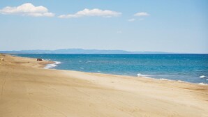Ein breiter Sandstrand mit sanften Wellen, blauem Himmel und fernen Bergen.