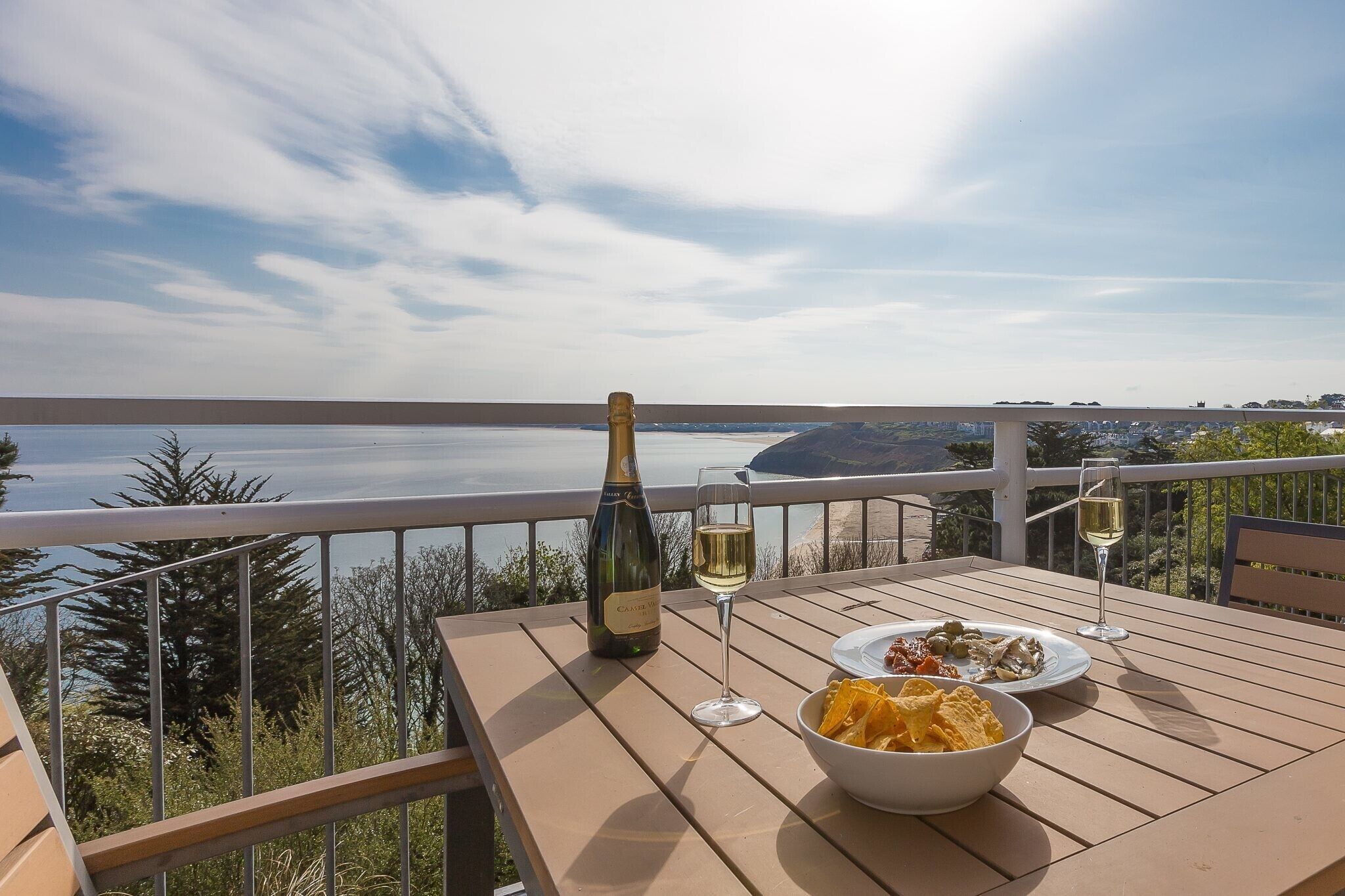 A table with a bottle of champagne, two glasses, and a bowl of chips on a balcony overlooking a body of water.