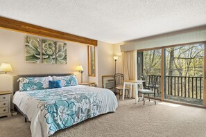 A bedroom with a large bed, a desk, and a balcony with a view of trees.