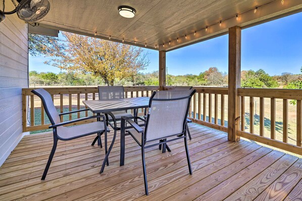 Une terrasse extérieure couverte, dotée d’une table et de chaises en bois, d’un ventilateur de plafond et offrant une vue sur le paysage environnant.