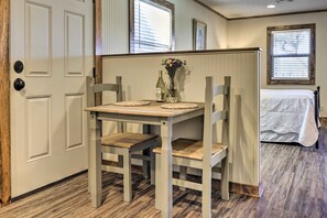 Une petite pièce agréable et chaleureuse, avec une table et des chaises en bois, un lit recouvert d’une couverture blanche et une fenêtre avec des stores.