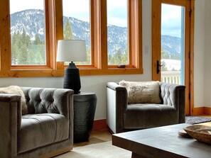 A room with two leather sofas, a wooden coffee table, and a lamp. Large windows with wooden frames offer a view of a mountainous landscape. - BRAND NEW LISTING! Gorgeous Private Big Sky Cabin! (Big Sky)
