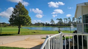 Vista de um campo de golfe com um lago, uma grande árvore e um edifício com janelas amplas.