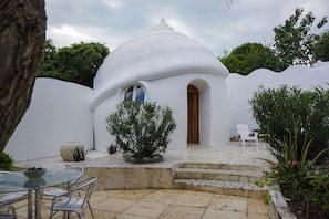A white dome-shaped building with a wooden door and windows, surrounded by greenery and a tiled patio area.