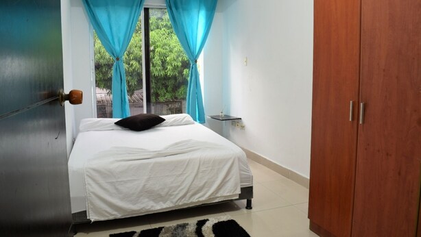 Un dormitorio con cama, un armario de madera, una ventana con cortinas y una mesita de noche con lámpara.