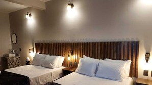 Quarto de hotel com duas camas, cada uma com lençóis brancos e cabeceira de madeira. Há luminárias laterais e uma pequena mesa com uma luminária em cada lado.