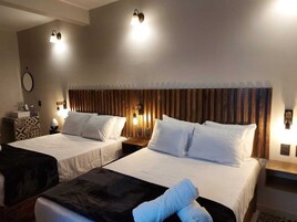 Quarto de hotel com duas camas, cada uma com lençóis brancos e cabeceira de madeira. Há luminárias laterais e uma pequena mesa com uma luminária em cada lado.