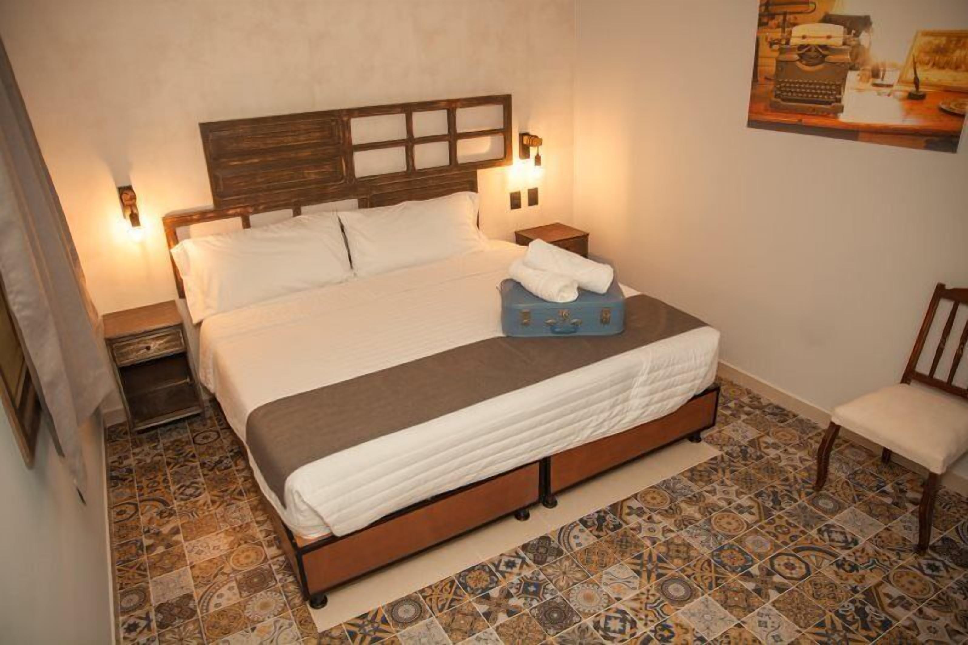 A bedroom with a wooden bed, white bedding, patterned floor tiles, a blue suitcase, and a chair.