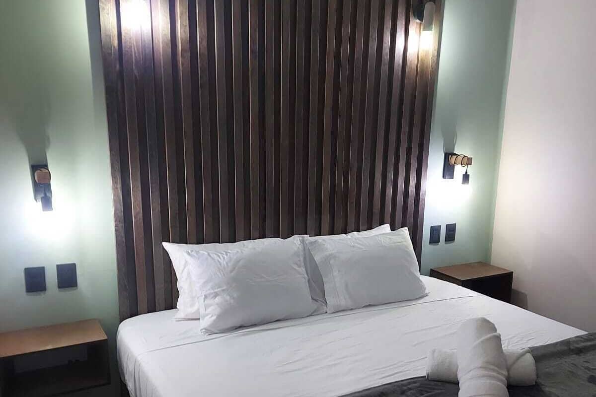 A hotel room with a large bed, two bedside tables, a headboard with vertical slats, and wall-mounted lighting.