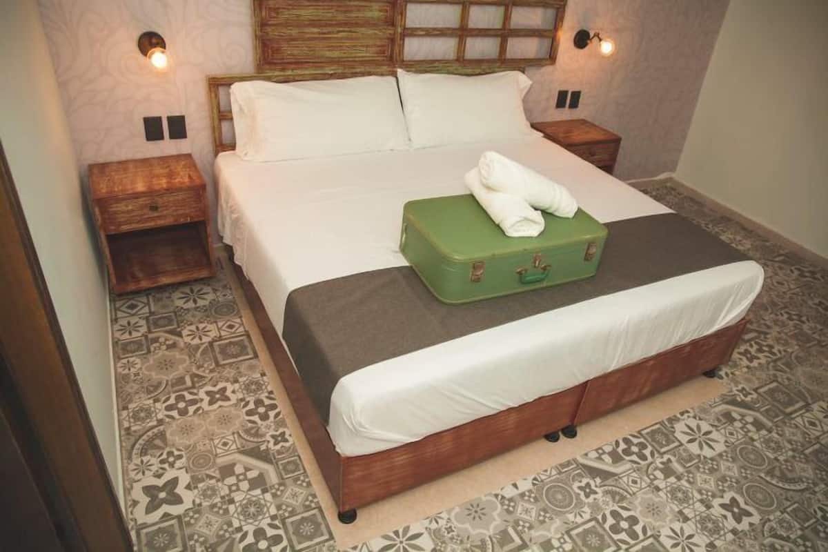 A neatly made bed with a green suitcase and a white towel on it, a wooden headboard, two bedside tables with lamps, and patterned floor tiles.