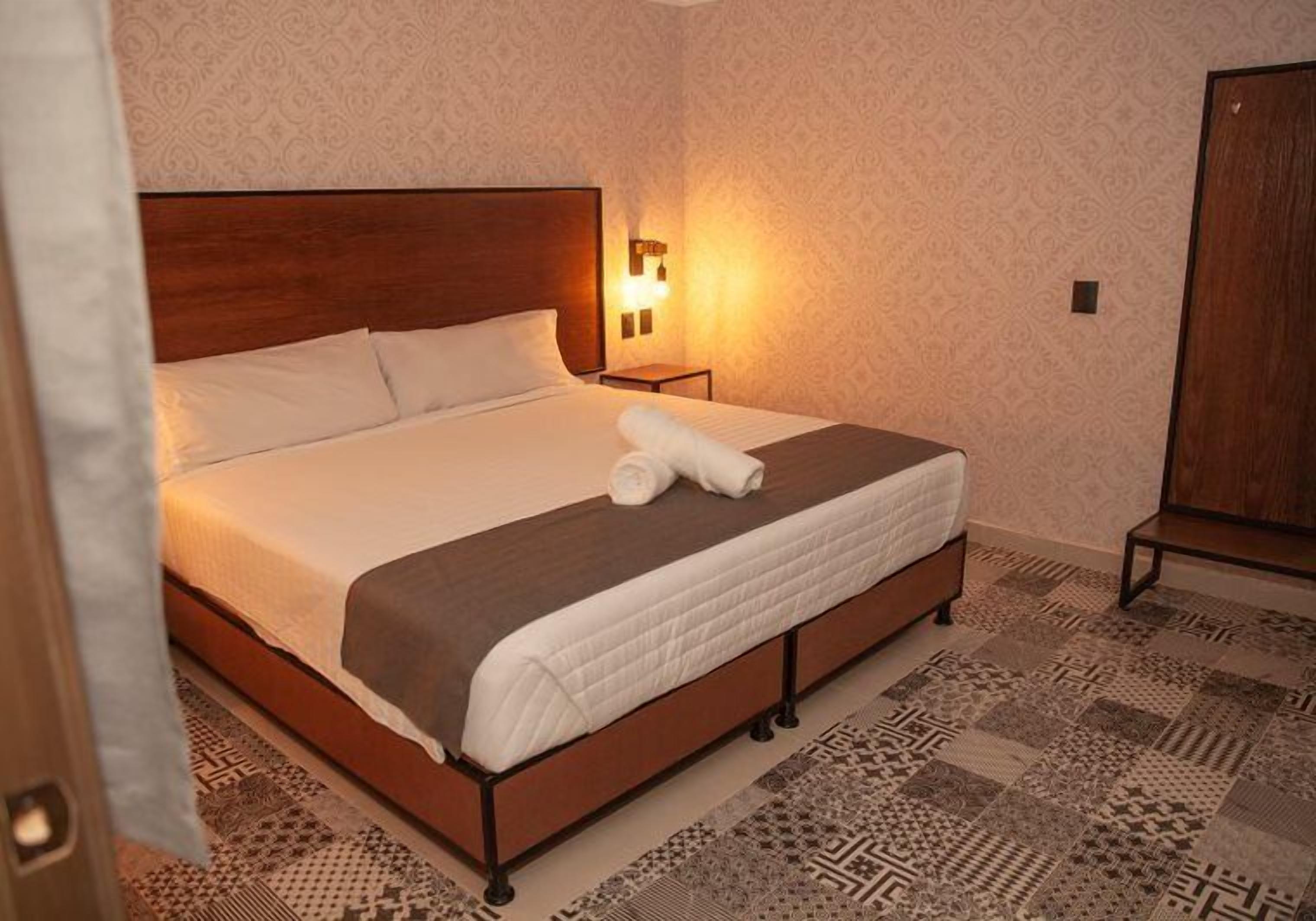 Een netjes opgemaakt bed met een bruin hoofdbord, een bedlampje en een kamerkleed op de vloer.