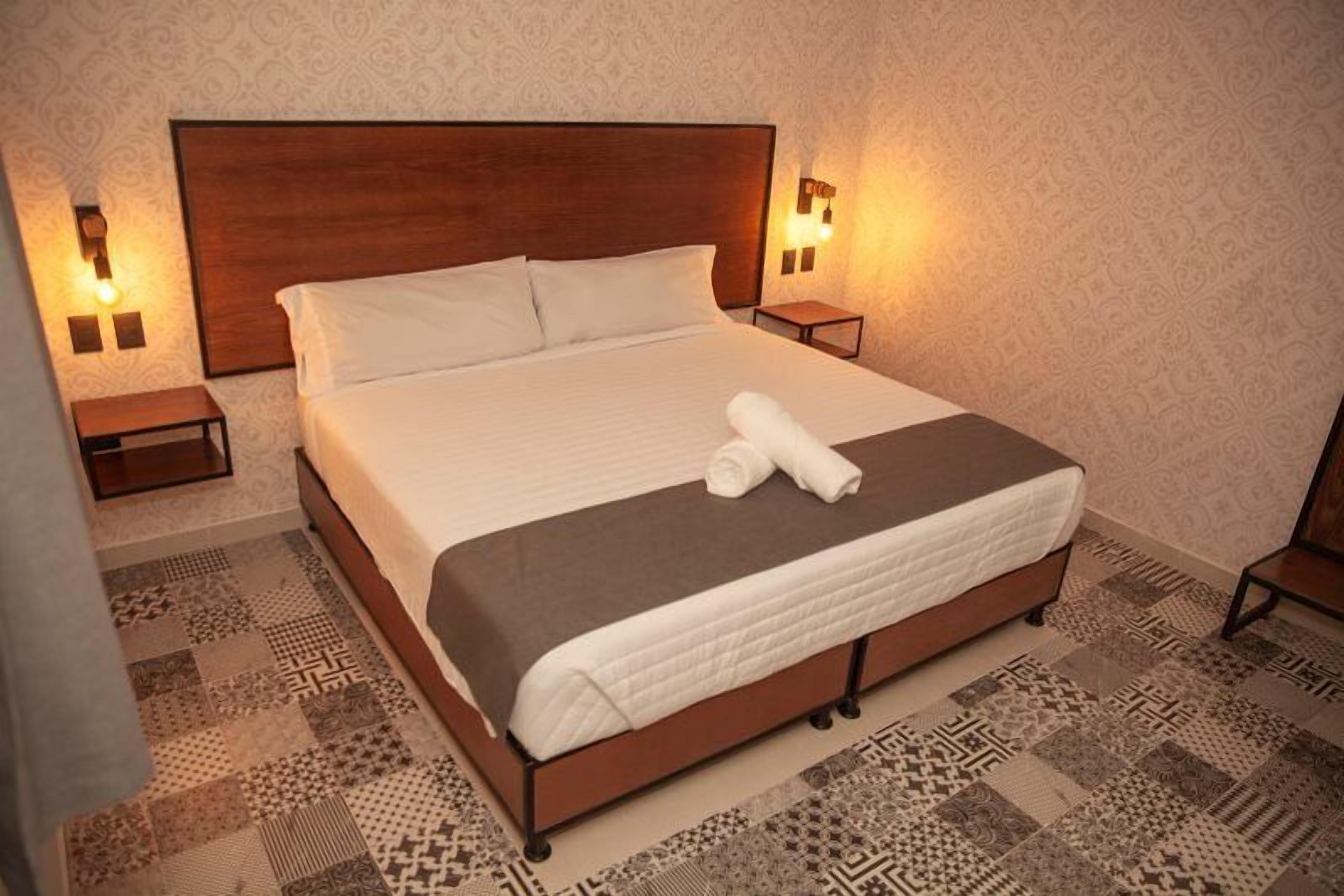 A neatly made bed with a brown headboard, two bedside tables with lamps, and patterned floor tiles.