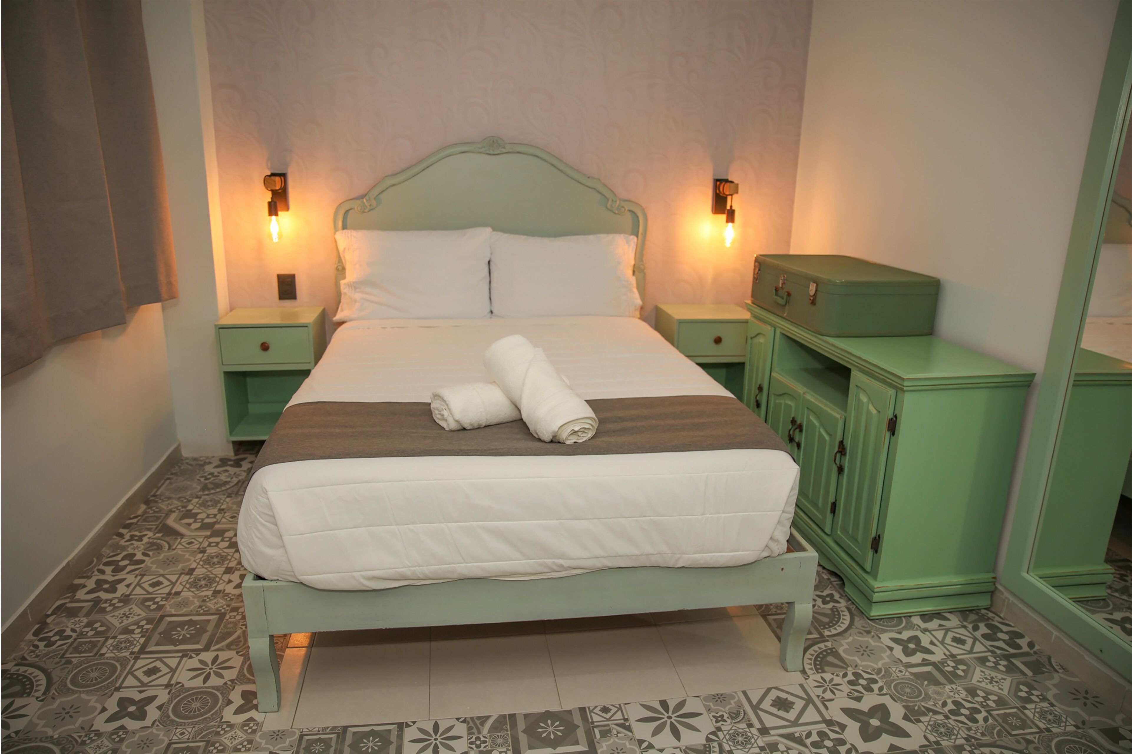 Una cama bien hecha con un edredón marrón y blanco, dos toallas enrolladas y una mesita de noche verde con una lámpara.