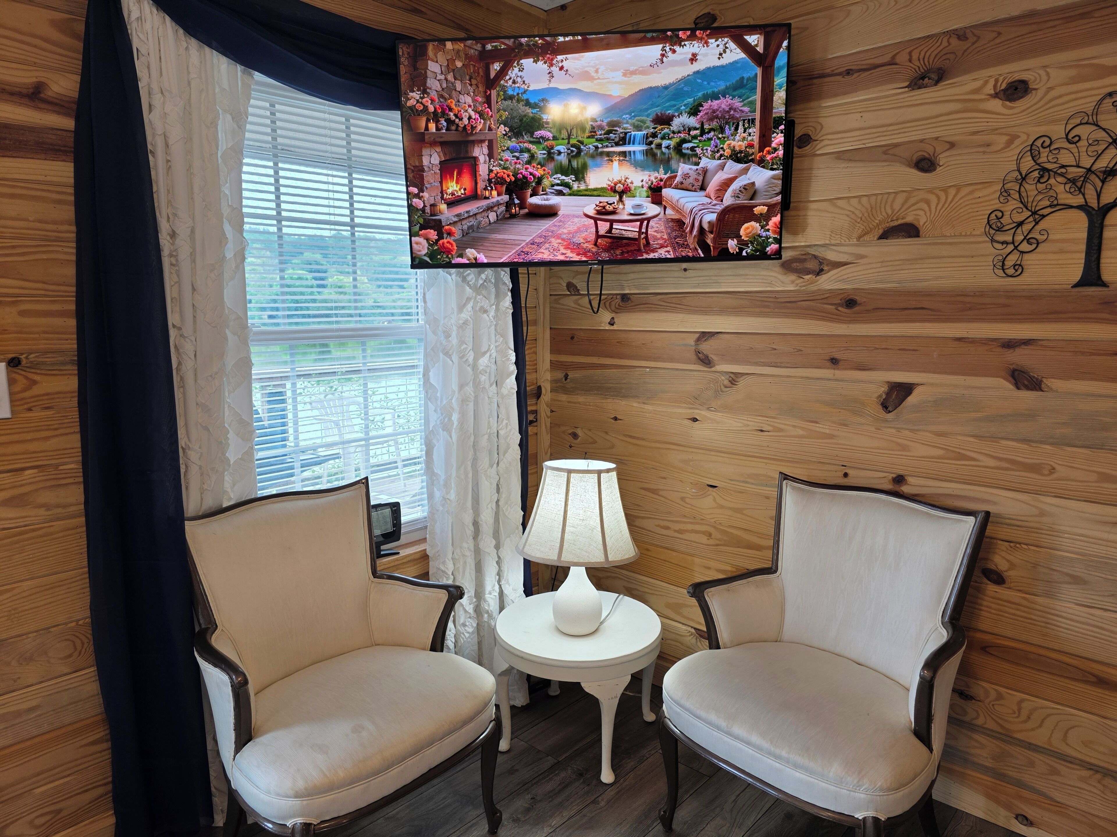 A cozy room with a TV mounted on the wall, two chairs, a small table with a lamp, and a window with curtains.
