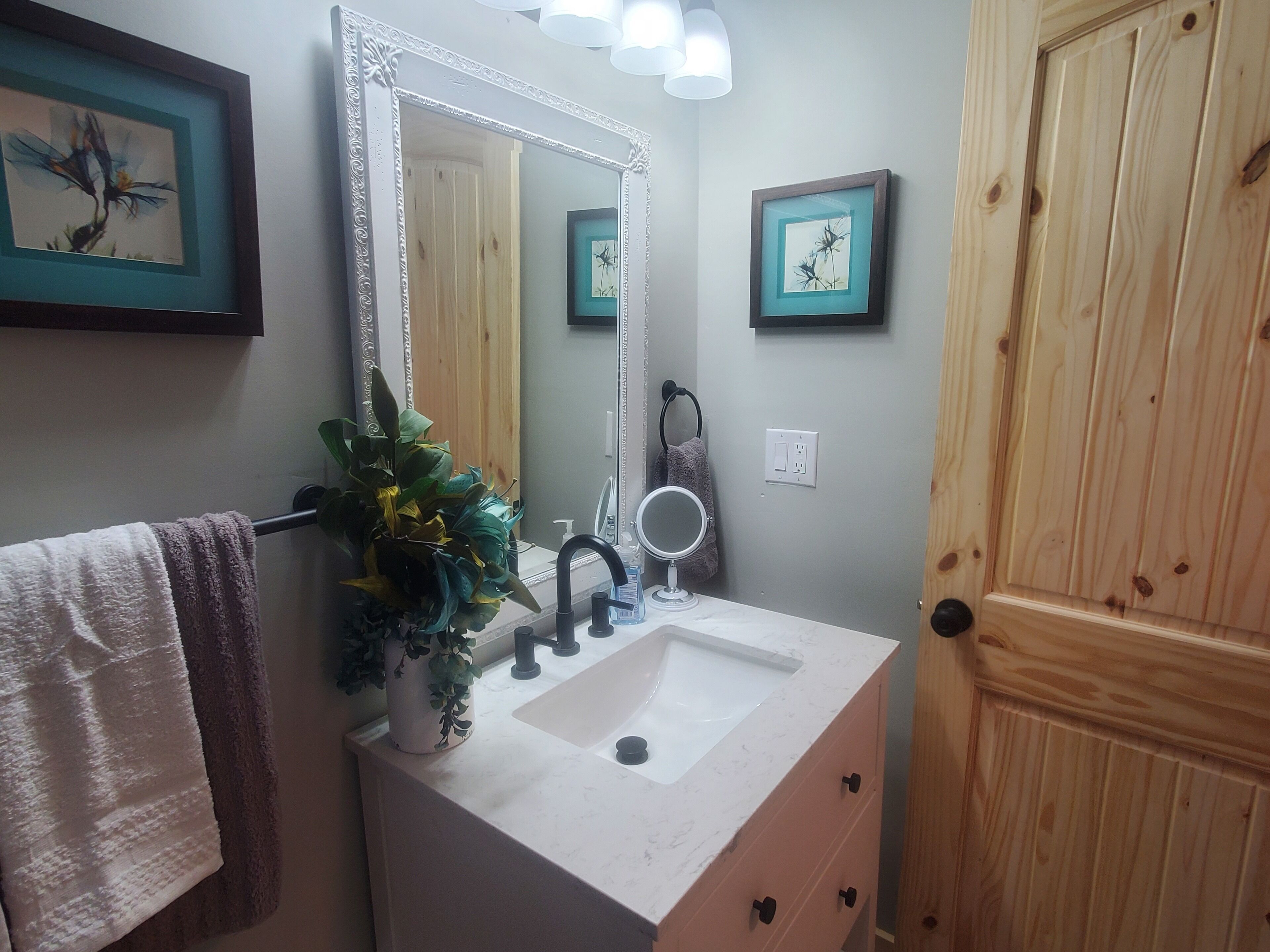 Un baño con lavamanos blanco, un espejo, una planta en maceta y puertas de madera.