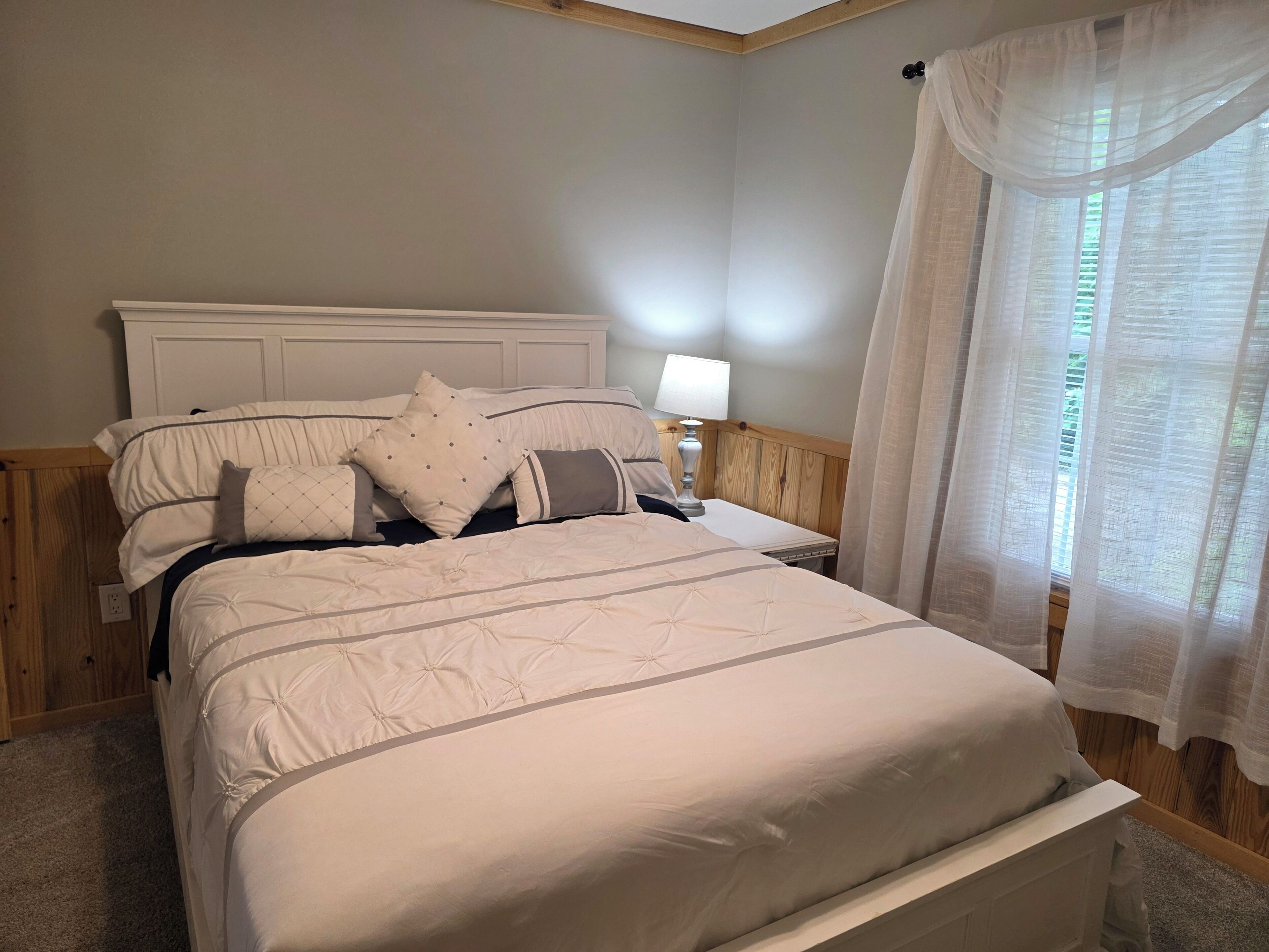 Uma cama com cabeceira, mesa de cabeceira com abajur e janela com cortinas translúcidas.