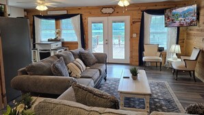 L’intérieur d’un chalet accueillant, avec un canapé, des fauteuils, une petite table, un téléviseur et une vue sur un lac, visible à travers la porte.