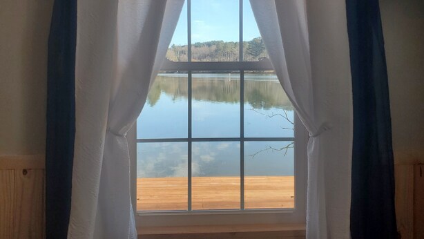 Une fenêtre avec des rideaux blancs, une vue sur un lac paisible et une terrasse en bois.
