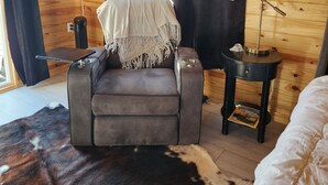 Une pièce chaleureuse avec un fauteuil en cuir, une petite table avec une lampe, un lit recouvert d’une couette blanche et un tapis en peau de vache.
