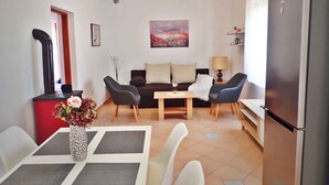 A compact living space with a dining area, a sofa set, a table with a vase of flowers, a painting on the wall, and a refrigerator.