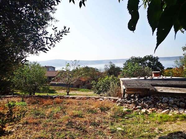 A rustic building with a stone foundation and a tiled roof, surrounded by a grassy area with trees and a view of the sea in the background.