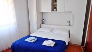 A neatly made bed with a blue bedspread and two folded white towels, a white wardrobe, and a small shelf with decorative items.