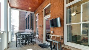 A covered outdoor seating area with a brick wall, a flat-screen TV mounted on the wall, and a table with chairs. - Majestic Lofts #302- Balcony over Printers Alley (Nashville)