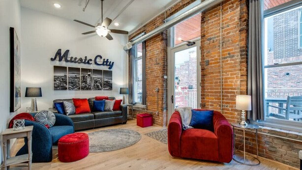A modern living room with a sofa set, red and blue chairs, a coffee table, and a large window with a city view. - Majestic Lofts #302- Balcony over Printers Alley (Nashville)