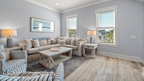 A spacious living room with a beige sectional sofa, a striped armchair, and a coffee table. There are two side tables with lamps, and large windows with blinds.