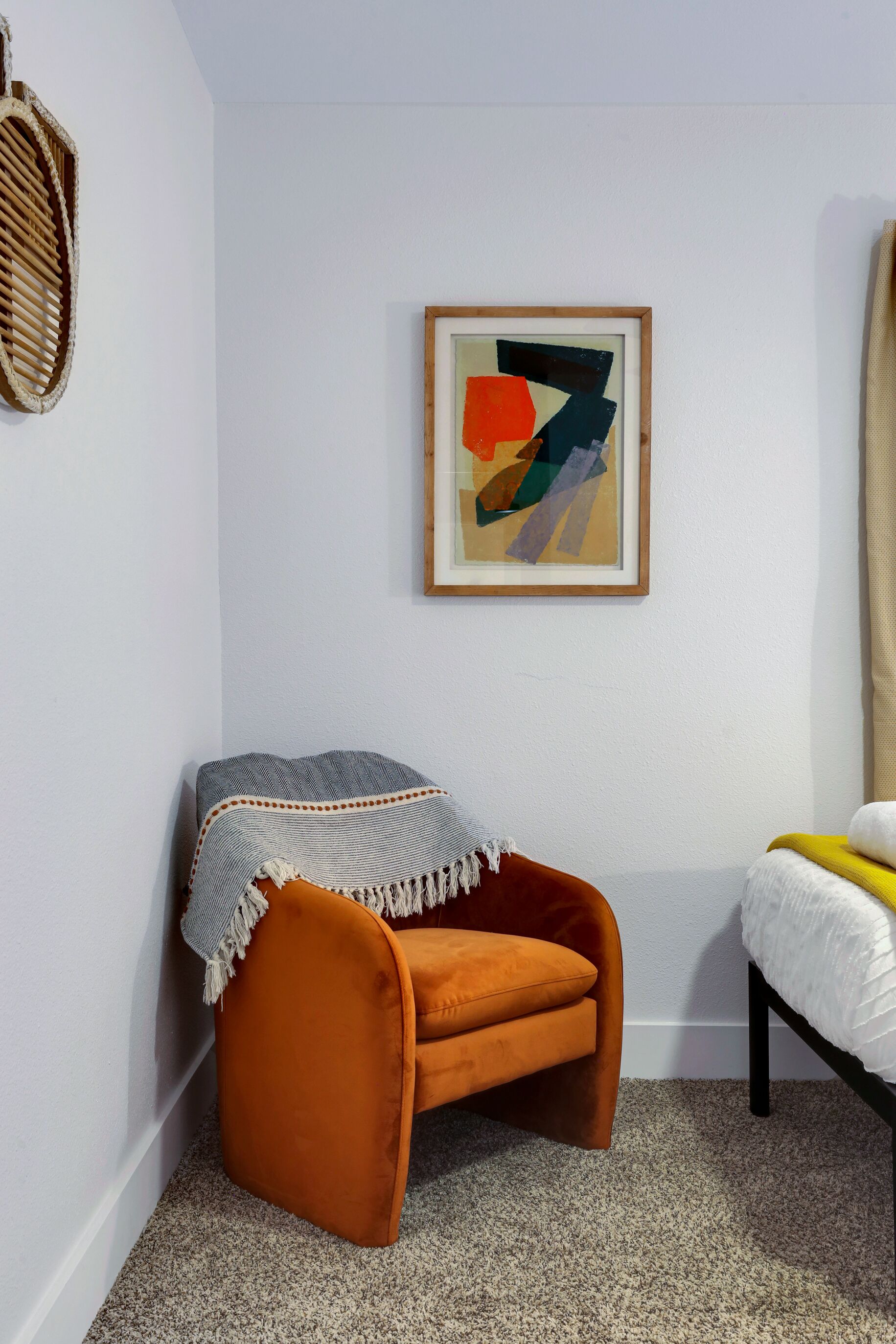 A corner room with a single orange chair, a framed abstract painting, and a bed with a white duvet.