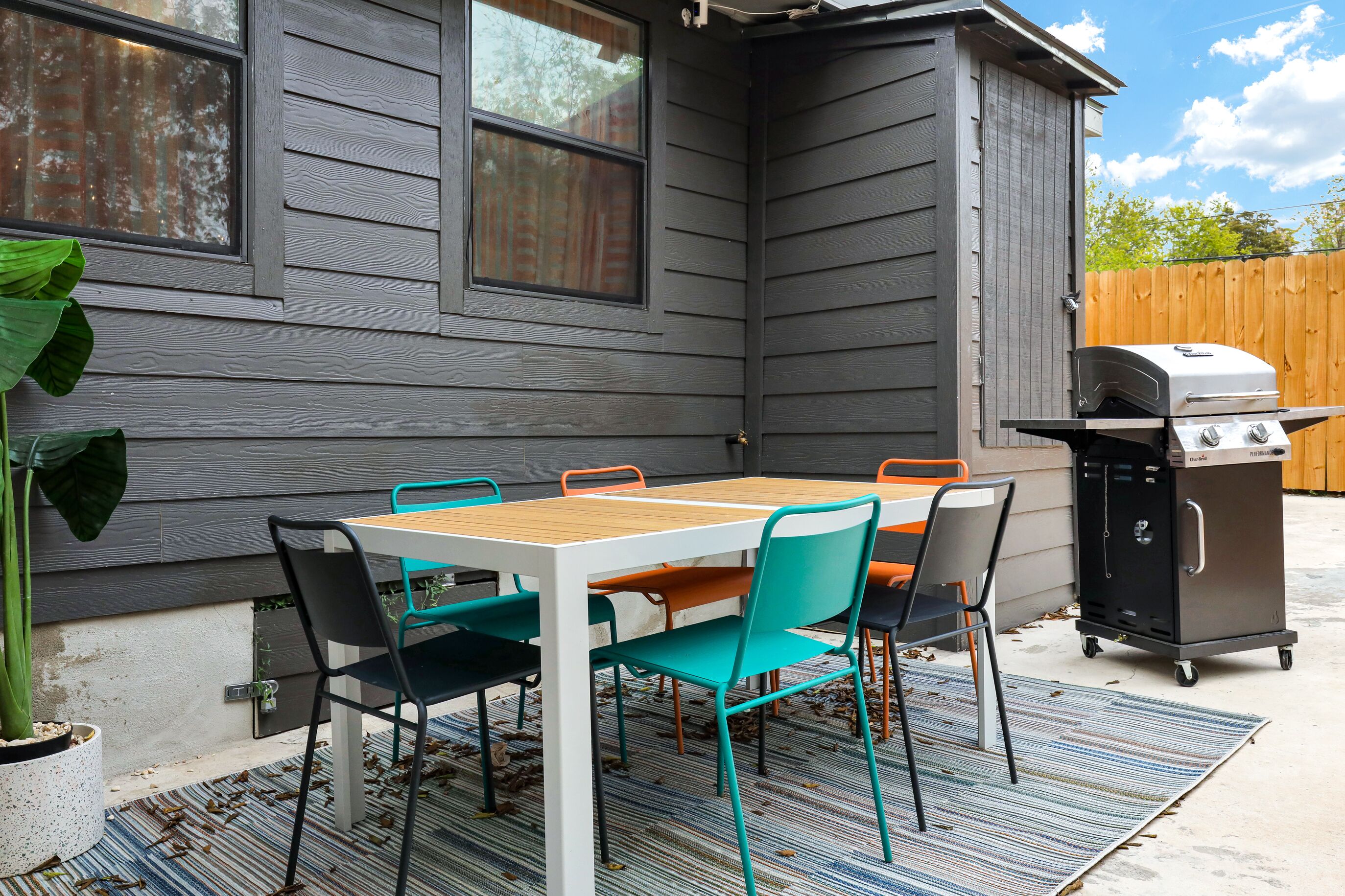 An outdoor patio with a wooden table and chairs, a grill, and a potted plant.