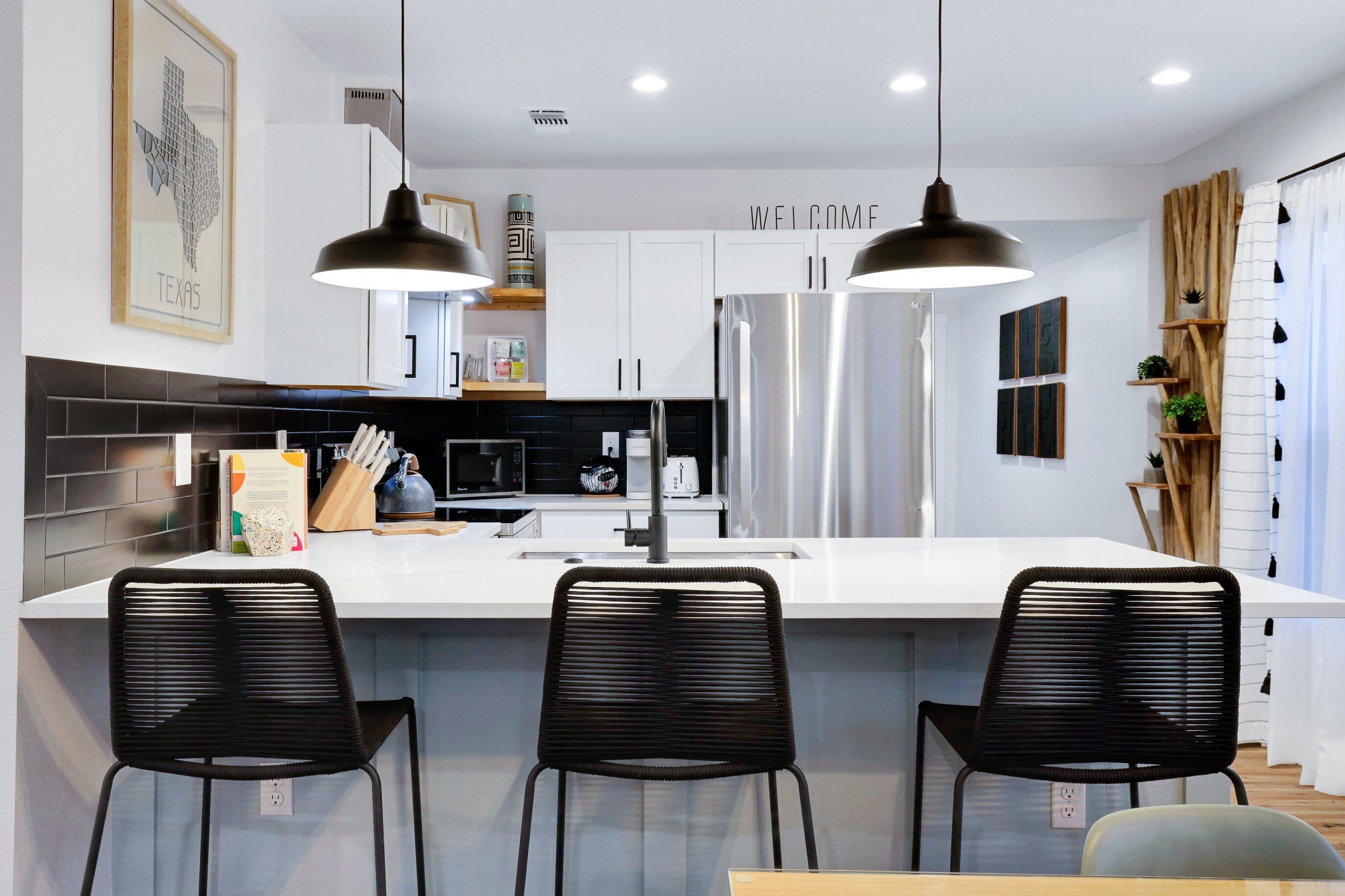 A modern kitchen with a central island, black chairs, stainless steel appliances, and a 'WELCOME' sign.