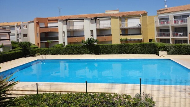 A swimming pool surrounded by apartment buildings with balconies, a clear blue sky, and a well-maintained hedge in the foreground. - Nice 2 room apt with Pool Richelieu beach Cap d'Agde (LE CAP D AGDE)