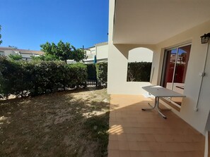 A patio area with a table and chairs, a small garden with a hedge, and a building with a window and door.
