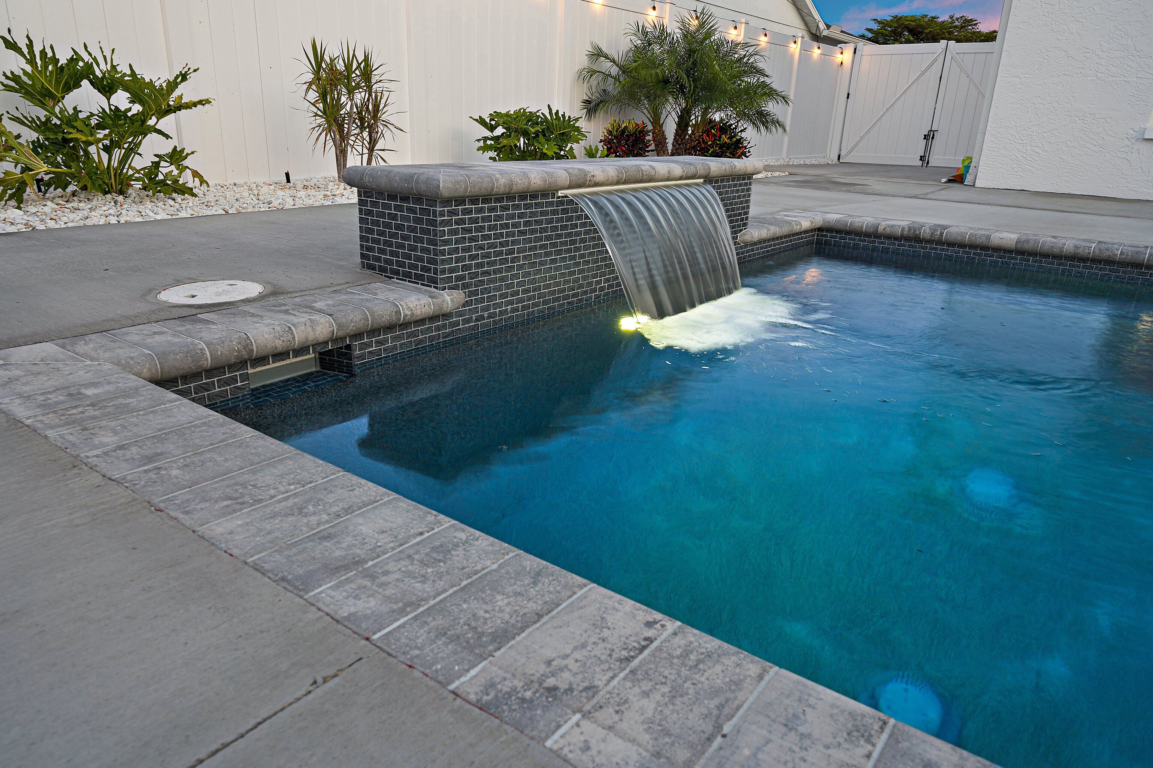 A swimming pool with a waterfall feature, surrounded by a tiled deck and various plants.